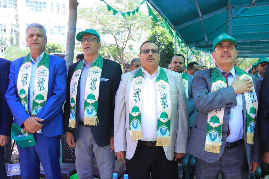 Group of men in suits wearing green ceremonial scarves, standing outdoors at a formal event under a tent.