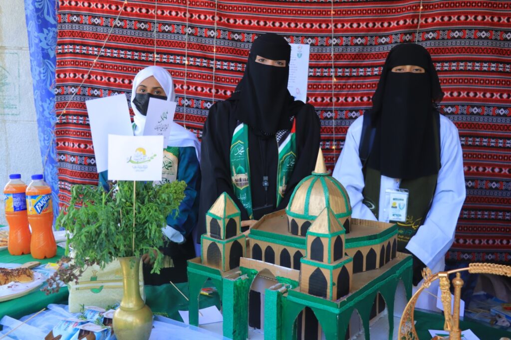 Three women in hijabs stand behind a table displaying a large green cardboard mosque model, herbs, and beverages against a red patterned backdrop.
