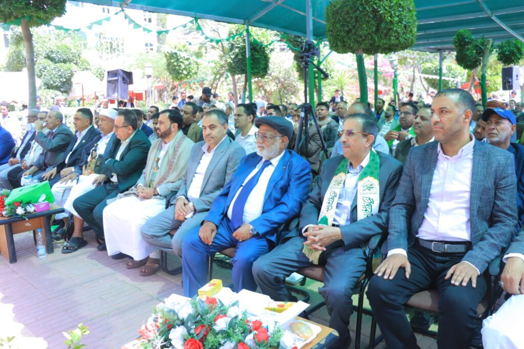 Group of formally dressed men seated outdoors under a teal canopy at a ceremony; foreground shows flowers and drinks on a table.