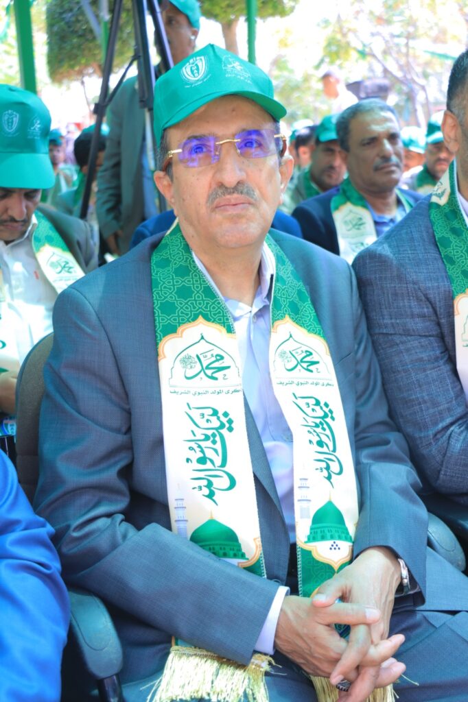 Man in a teal cap and matching scarf seated at an outdoor event, hands folded, facing forward amidst a group of people wearing similar attire.