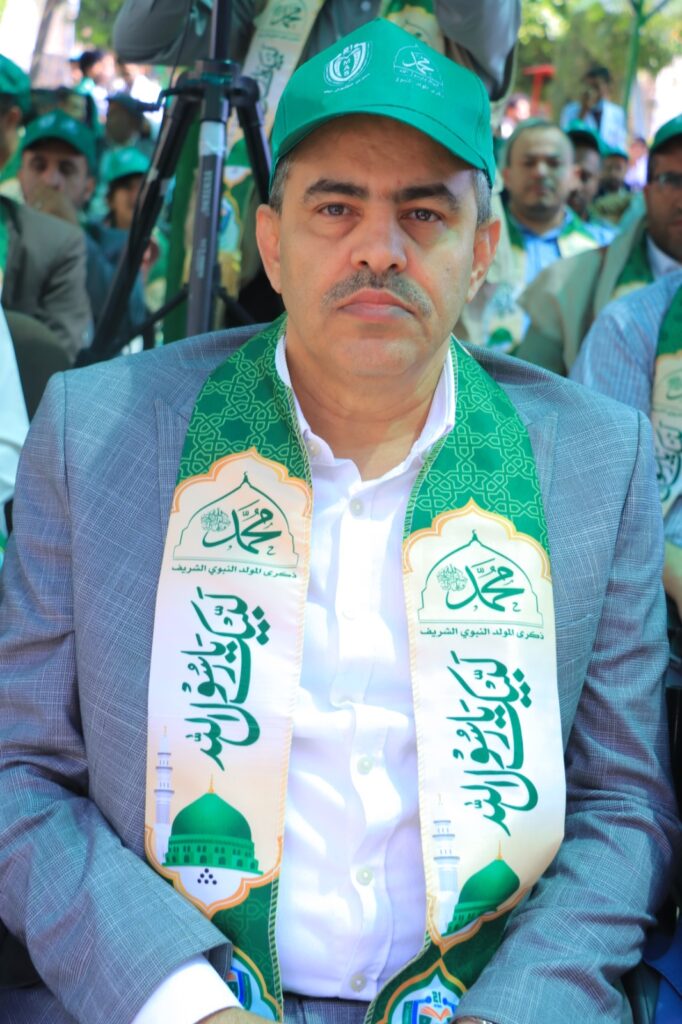 Middle-aged man in a gray suit with a white shirt, wearing a green scarf and cap at an outdoor ceremony. A crowd sits behind him.