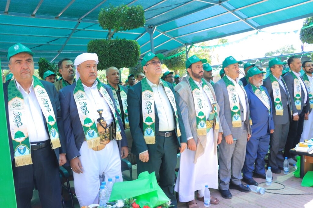 Group of men in suits wearing green and white scarves and green caps at an outdoor ceremony under a teal canopy.