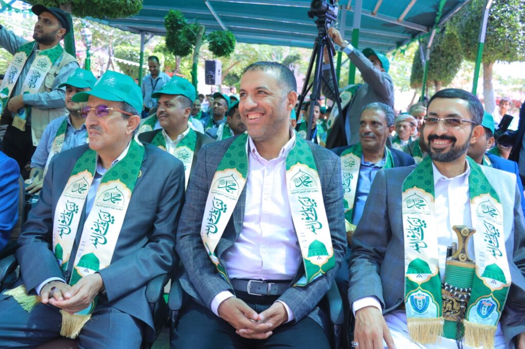 Group of men in suits wearing green ceremonial sashes at an outdoor event, smiling and seated under a canopy.