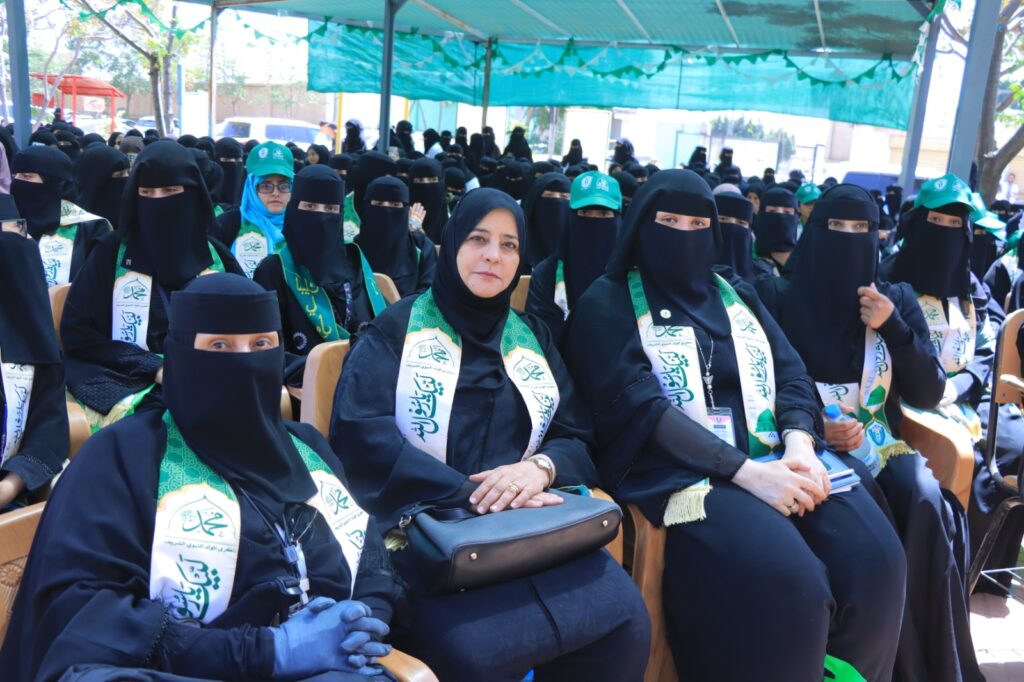 Group of women in black abayas and niqabs seated at an outdoor ceremony under a green canopy, wearing green and white sashes.