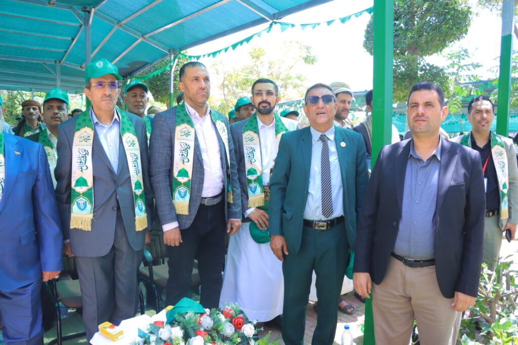 Group of men in suits and traditional attire wearing green scarves at an outdoor ceremony under a teal canopy, with green bunting in the background.