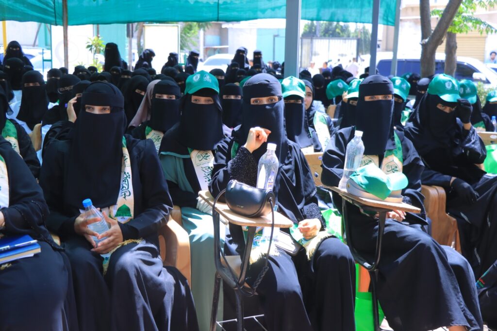 Group of women in black abayas and niqabs seated outdoors, many wearing green caps and sashes at a ceremony.