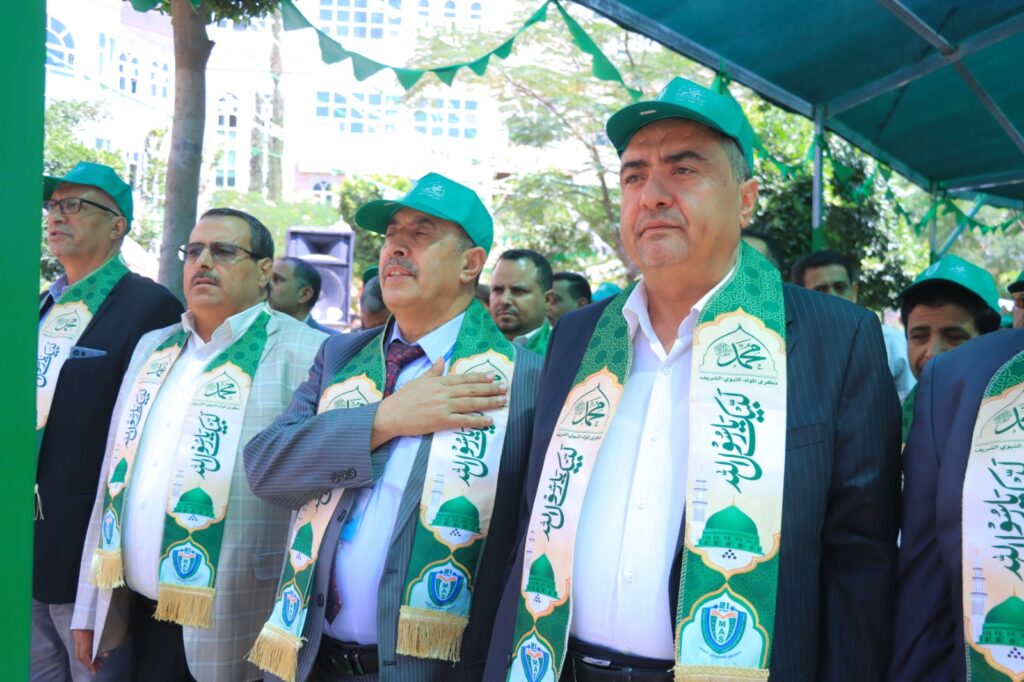 Group of men in suits wearing green ceremonial scarves and caps at an outdoor event, one man placing his hand over his heart.