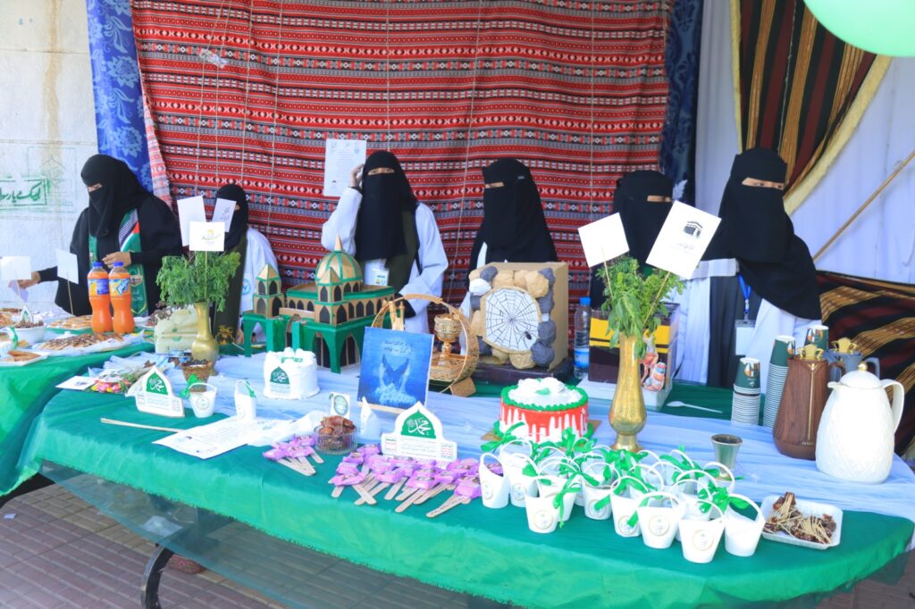Six women wearing niqabs stand behind a long table at a bazaar stall, displaying handmade crafts, cakes, drinks, and plants against a red patterned backdrop.