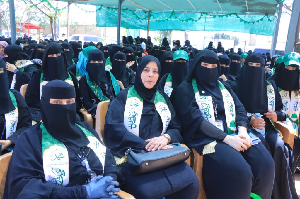 Large crowd of women wearing black abayas and niqabs seated at an outdoor event, each with green sashes under a teal shade canopy.