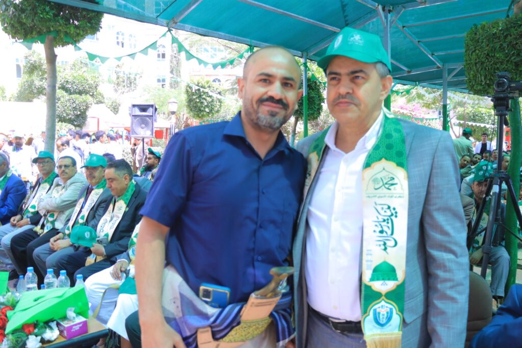 Two men posing at an outdoor ceremony under a teal canopy; one in a blue shirt, the other in a suit with a green ceremonial sash.