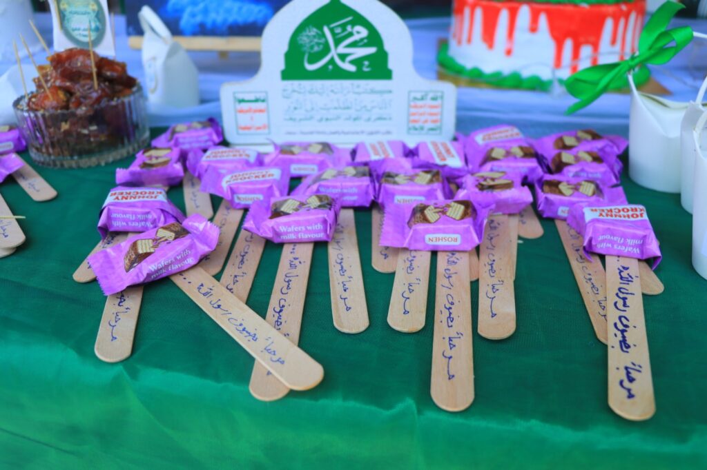 Assorted purple-wrapped wafer candies on wooden sticks laid out on a green table at a celebration, with a cake in the background.