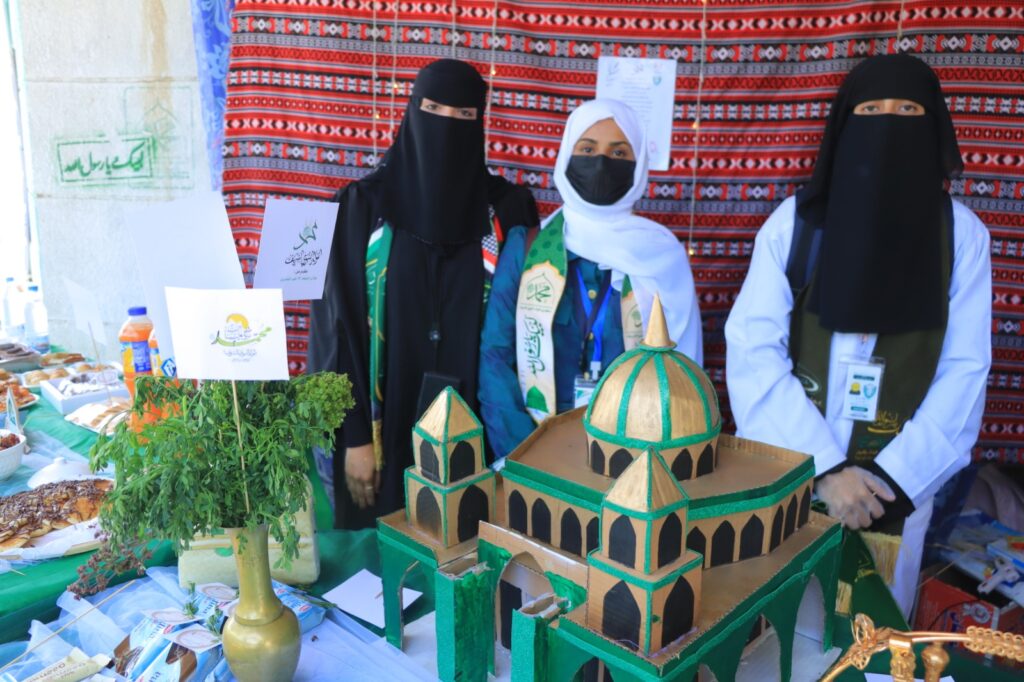 Three women wearing hijabs/niqabs stand behind a food stall with a green and gold cardboard mosque model in front of them and a patterned backdrop.
