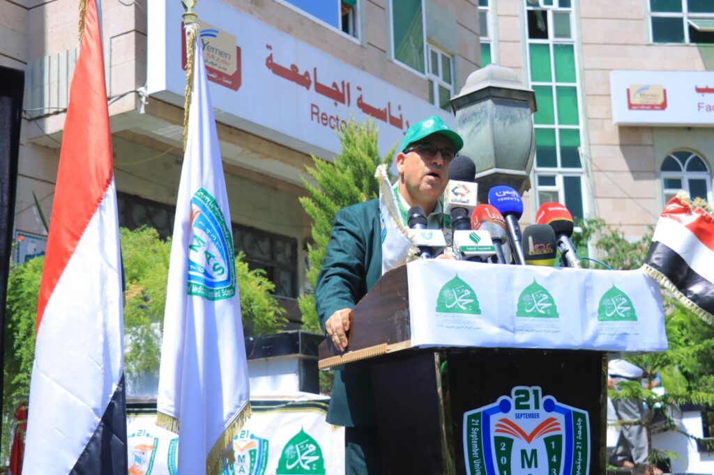 Speaker at a podium during an outdoor event, surrounded by microphones, banners, and Yemeni flags.