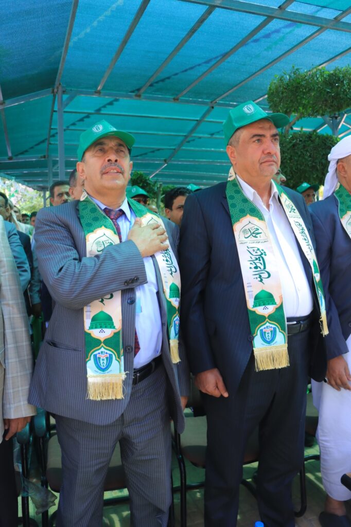 Two men in suits wearing green caps and ceremonial green scarves stand at an outdoor event under a blue canopy.