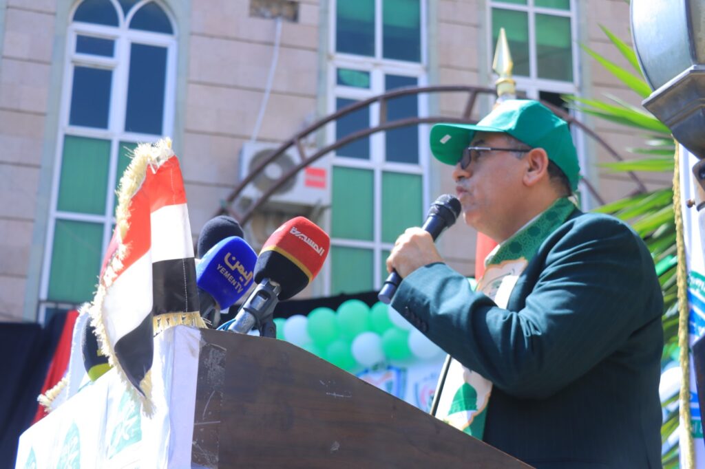 Man in green cap and suit speaks into a microphone at a podium during an outdoor event, with microphones and flags nearby.