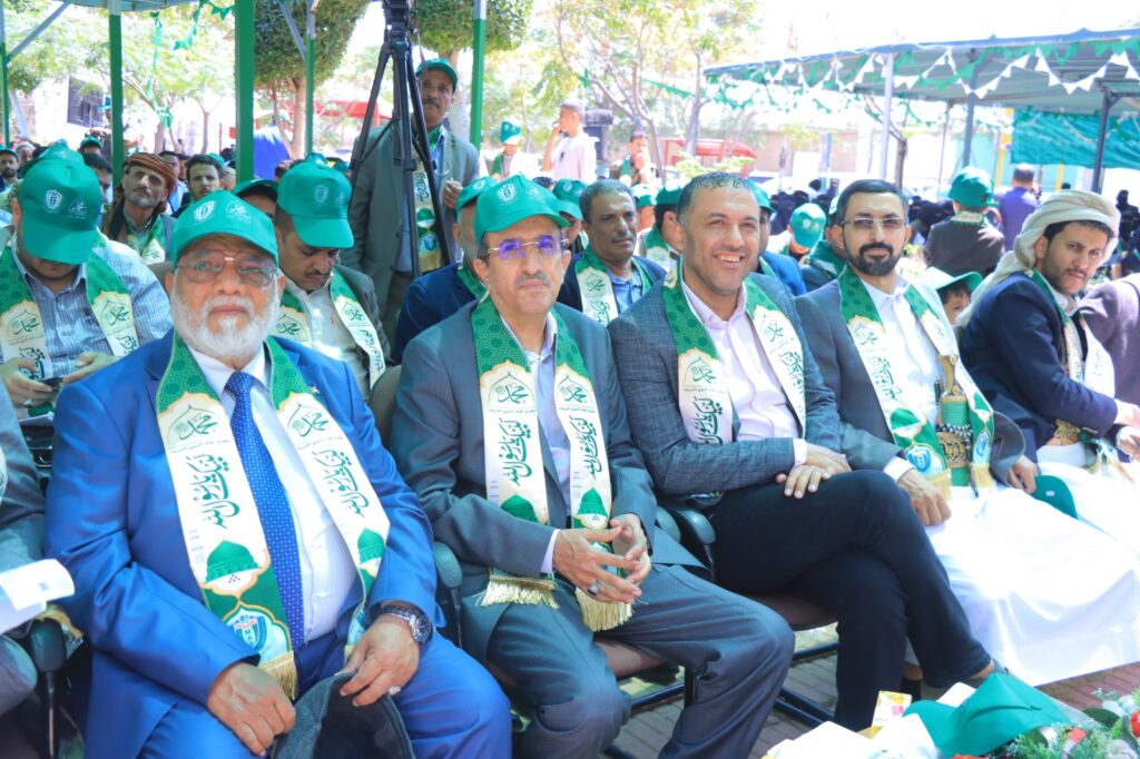 Group of men in suits wearing green hats and ceremonial sashes at an outdoor event under a canopy.