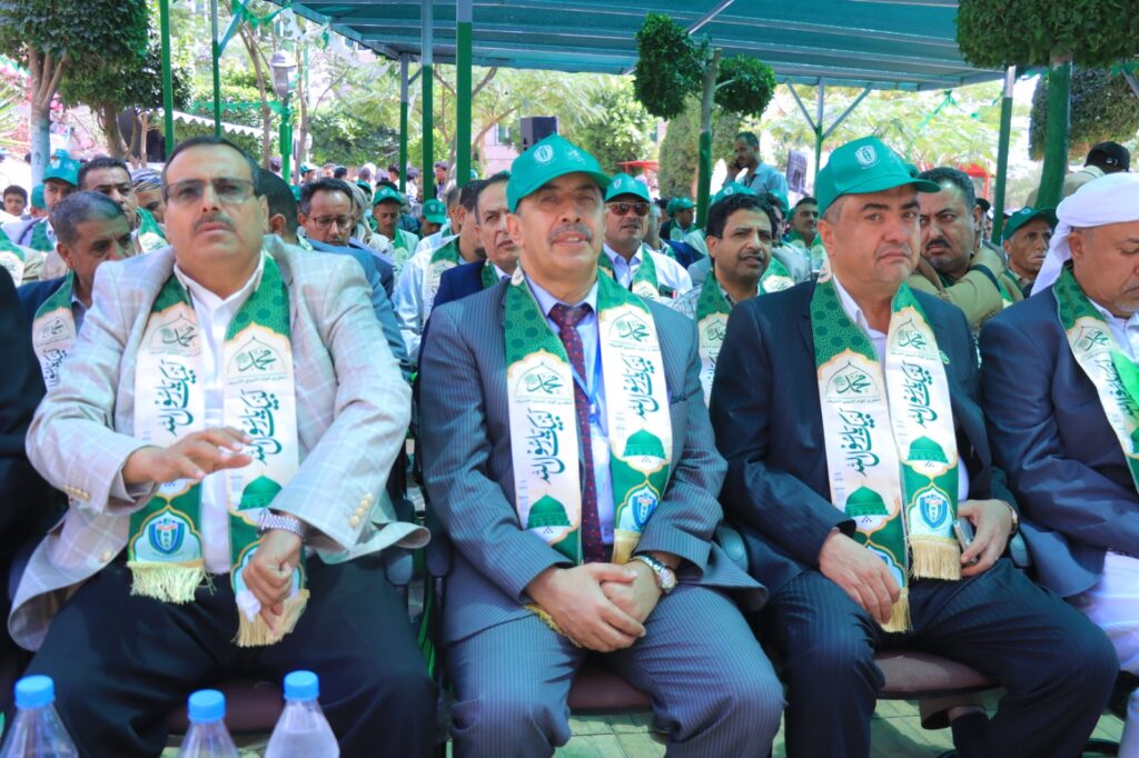 Group of men in suits seated at an outdoor ceremony, wearing green scarves with logos and matching green hats under a canopy.