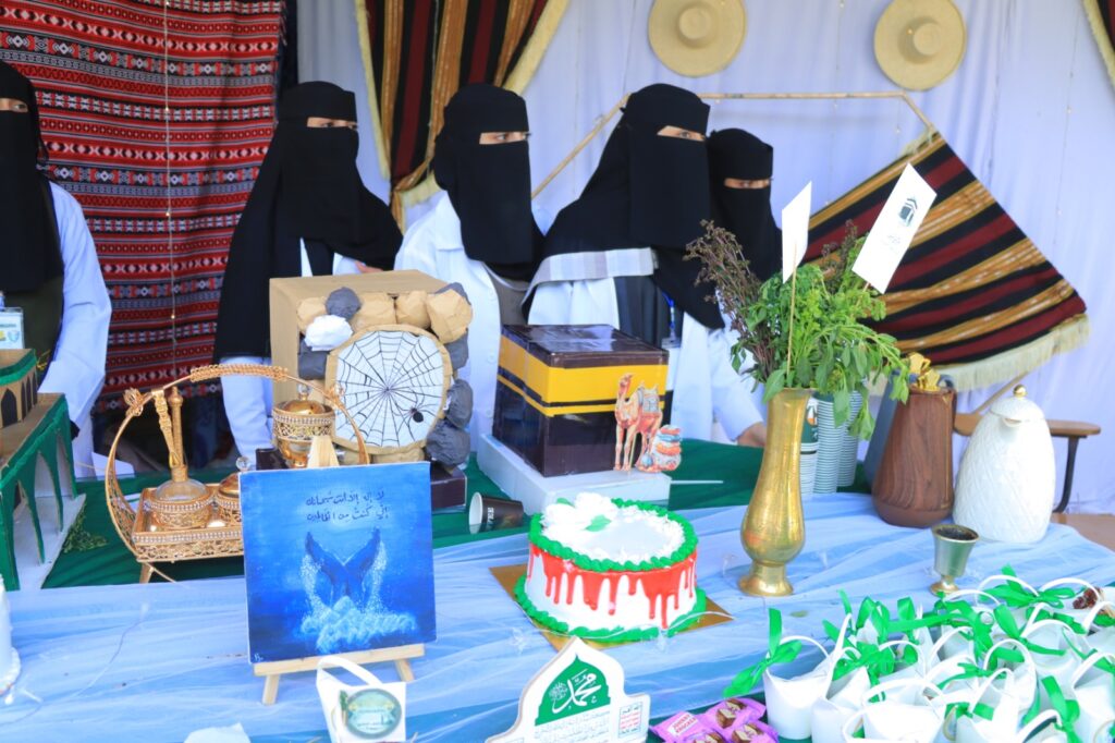Group of women in black niqabs standing behind a display table with cakes, crafts, and colorful fabrics at a cultural fair.