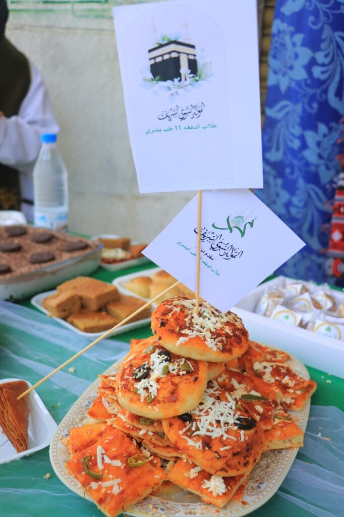 Stack of orange-topped savory pastries arranged on a decorative plate at an outdoor market stall, with white flags bearing Arabic script above them.