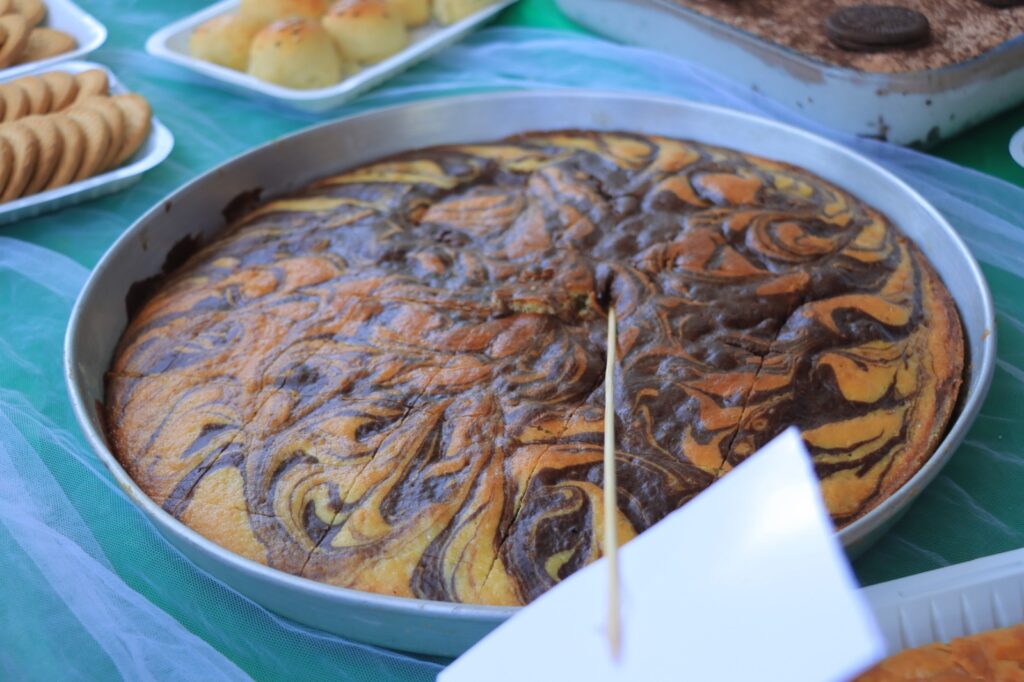 Round baking pan with a marble swirl cake (chocolate and vanilla) on a table with cookies and pastries in the background.