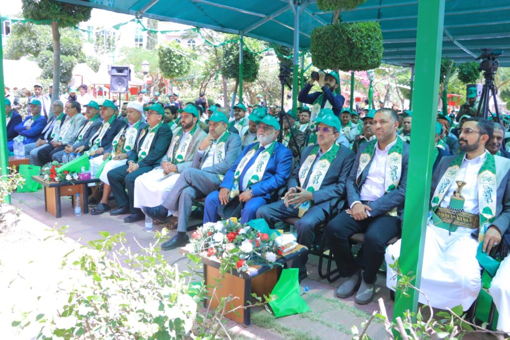Outdoor formal ceremony: a long row of men in suits with green scarves and hats seated under a green canopy, with photographers and greenery in the background.