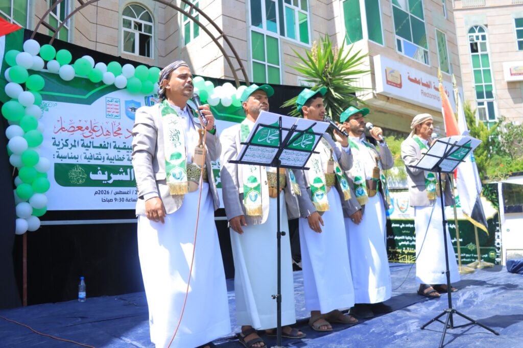 Group of men in white robes and green scarves performing on a stage with microphones, in front of a banner and balloons outside a building.