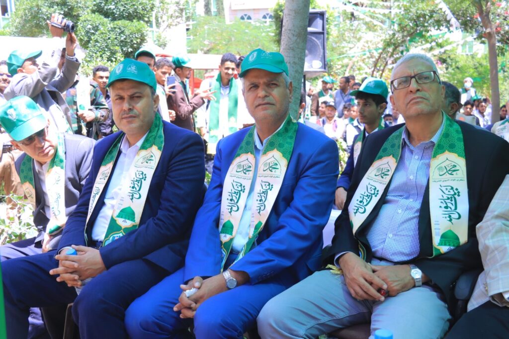 Men in blue suits with green caps and scarves seated at an outdoor event, crowd in background under leafy trees.