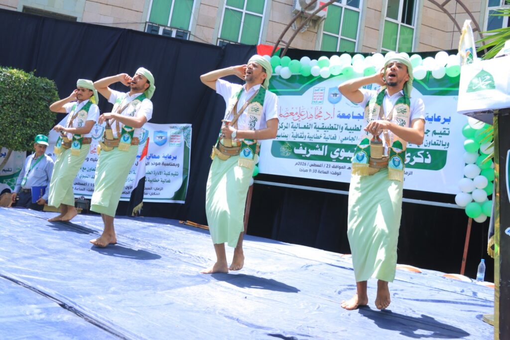 Four men in matching light-green traditional outfits perform a coordinated salute on a blue stage, barefoot, with a green-and-white balloon backdrop and Arabic banners in the background.