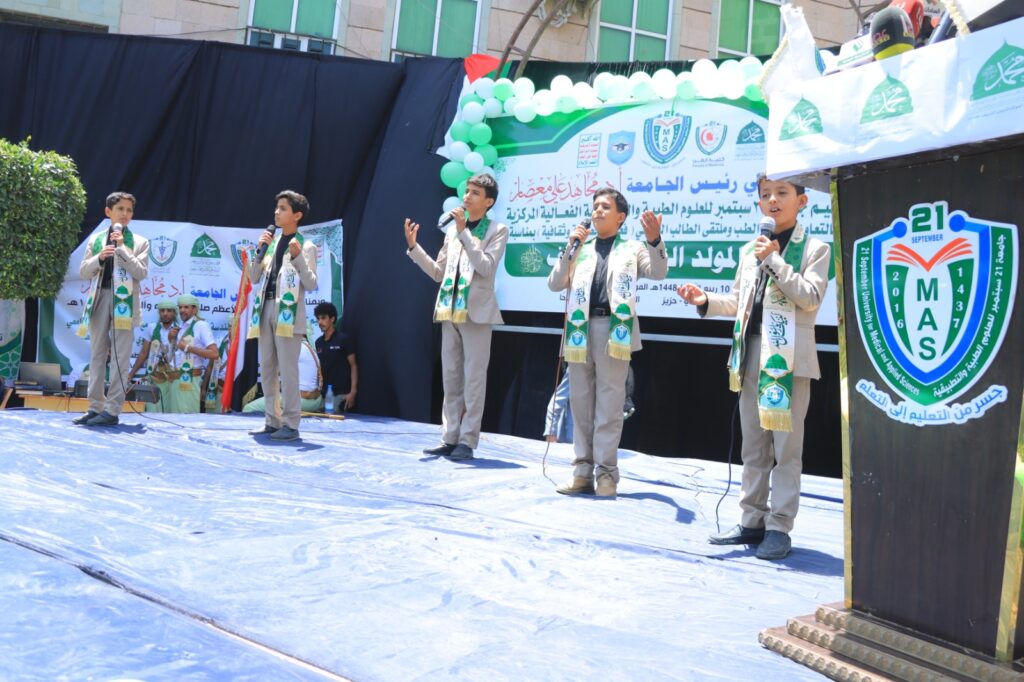 Six boys in light suits sing into wireless microphones on a stage during a school event, green scarves hanging over their shoulders with a large banner and MAS logos in the background.