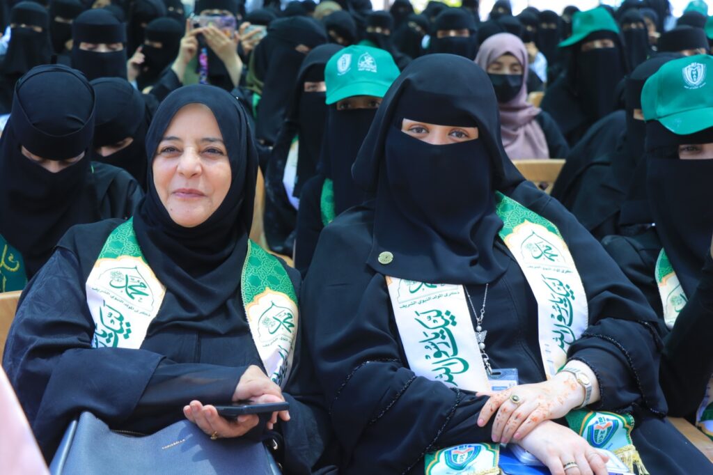 Two women in black hijabs with green ceremonial sashes sit in the front row at a formal event, smiling at the camera.