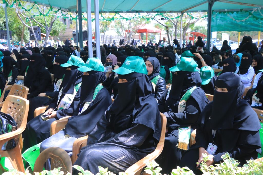 Group of women in black abayas and niqabs seated at an outdoor event, many wearing teal caps and sashes for a ceremony.