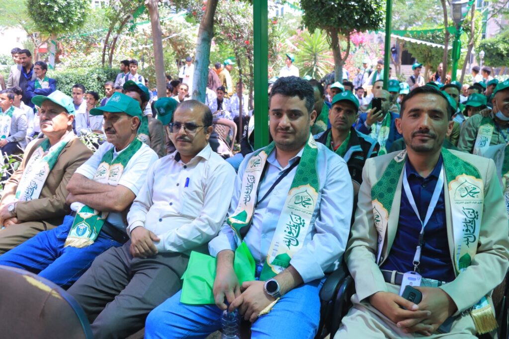 Group of men wearing green scarves and caps seated in a row at an outdoor gathering, with a crowd and trees in the background.