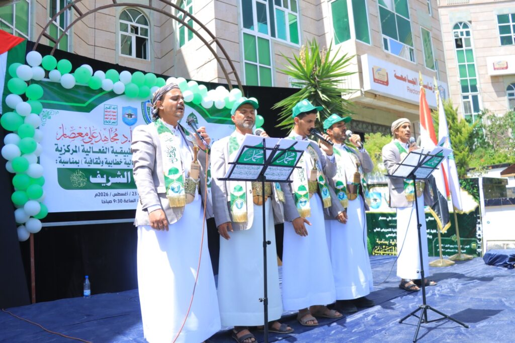 Group of men in traditional white robes singing on a stage with microphones, banners, and green balloons at an outdoor event.