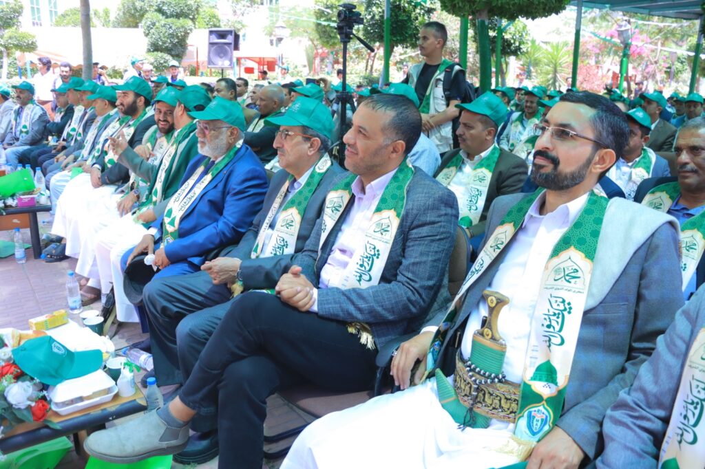 Men in green scarves and caps sit in rows at an outdoor ceremony; a camera on a tripod behind them.