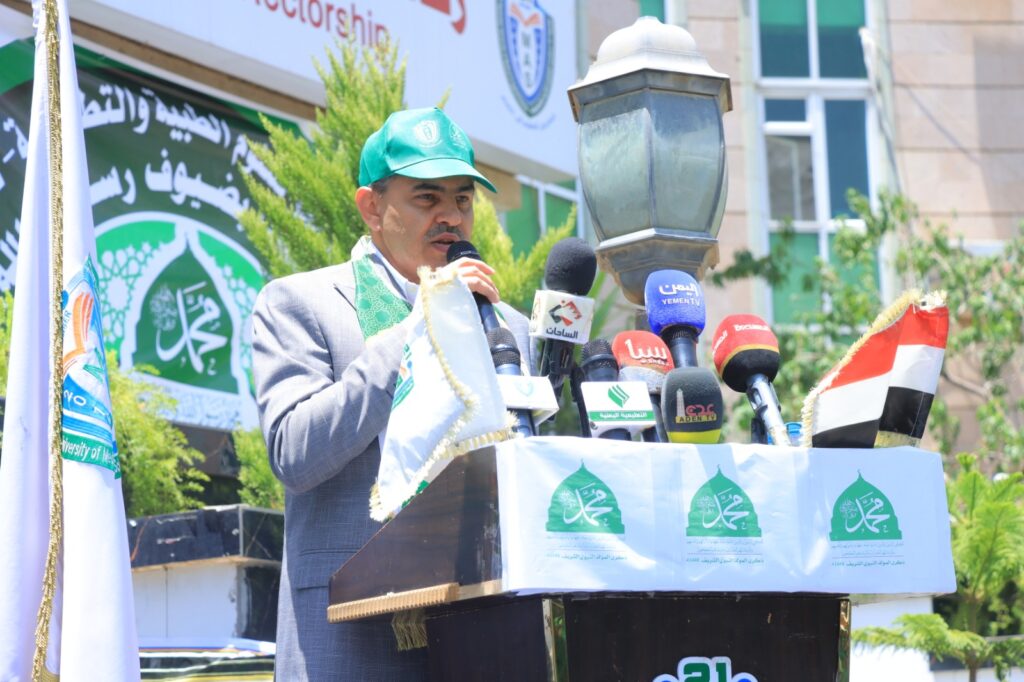 Man in a suit and green cap speaking into a microphone at an outdoor event, surrounded by news microphones at a podium with banners in the background.