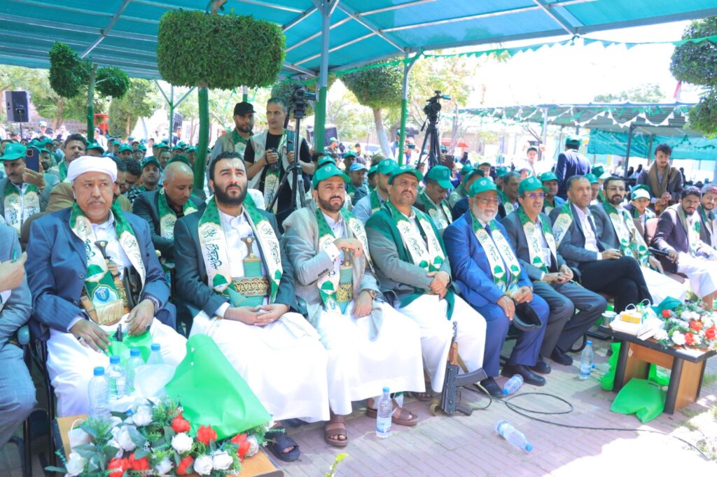 Group of men in traditional attire with green scarves seated at an outdoor ceremony under a turquoise canopy; photographers and a crowd in the background.