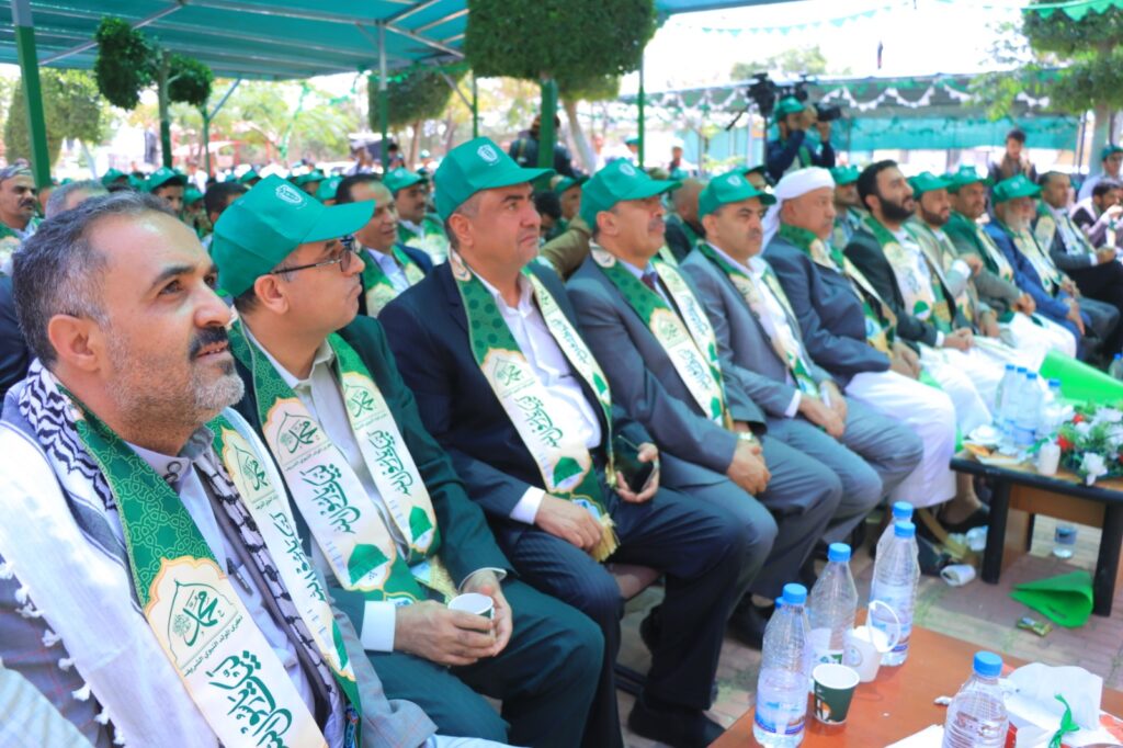Group of men in suits wearing green caps and matching sashes at an outdoor ceremony, seated under turquoise canopies with tables of water bottles and cups nearby.