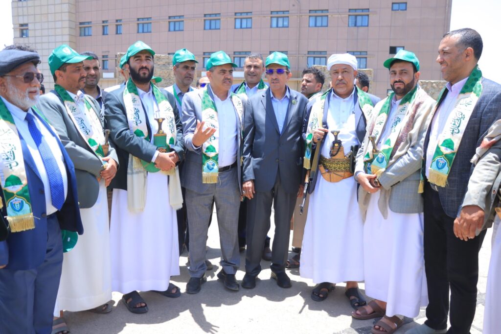 Group of men wearing green sashes and caps in a ceremony outdoors, some in traditional white robes and others in suits, standing together near a modern building.
