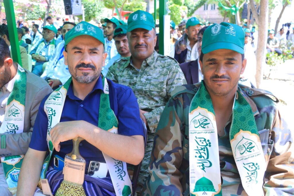 Two men in green caps and ceremonial scarves sit at an outdoor event, with a man in camouflage behind them and a crowd in the background.