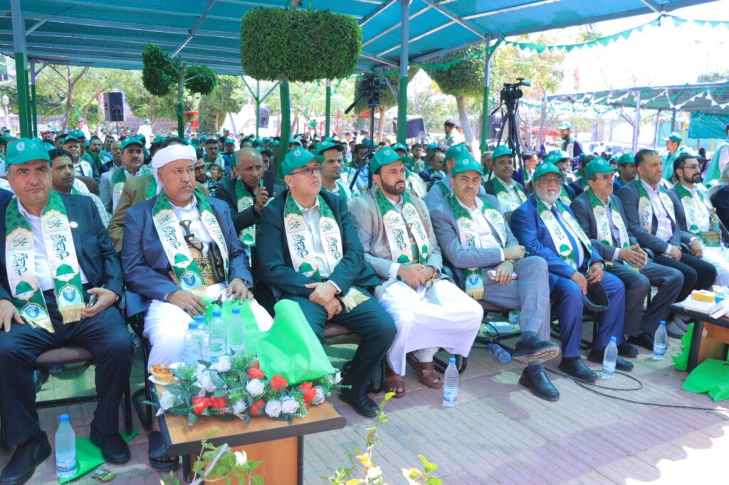 Group of men in green caps and sashes seated at an outdoor ceremony under a blue canopy with a crowd in the background to the side and cameras nearby.