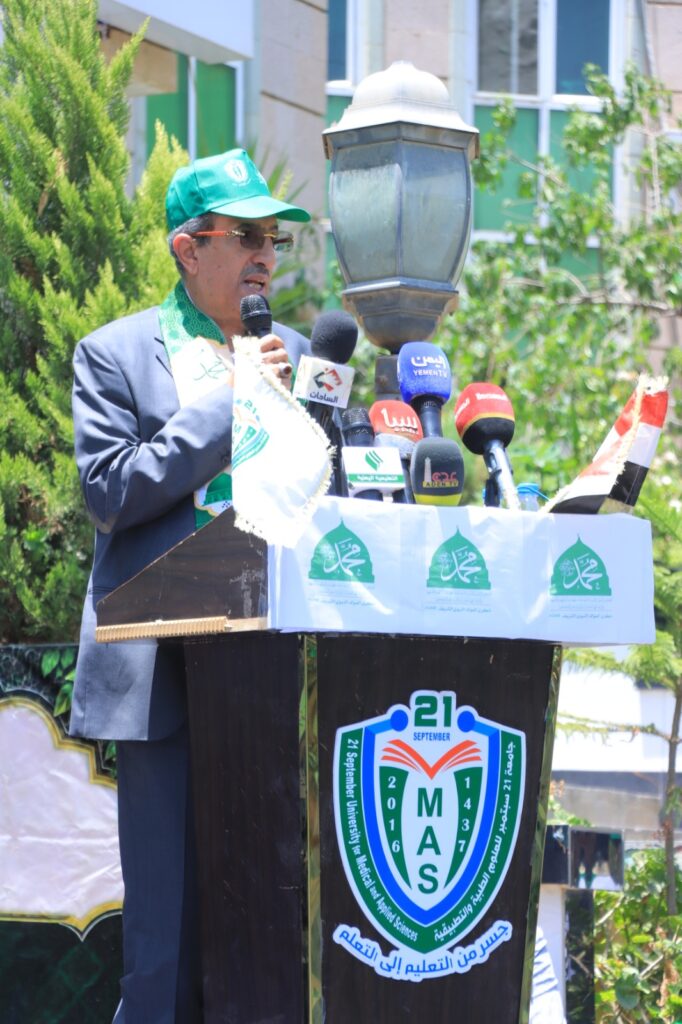 Man in a suit, green cap and scarf, speaks into a microphone at a podium with MAS logos and numerous microphones outdoors.