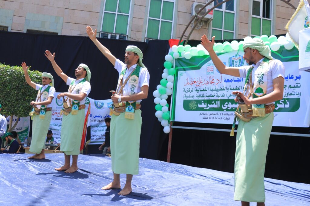 Four men in white shirts, green sashes and turbans perform a traditional dance on an outdoor stage, holding curved swords with raised arms on a blue floor.