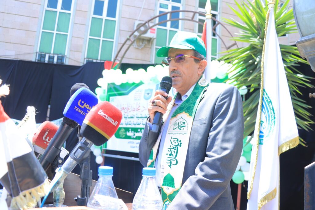 Man in a suit and green cap speaks into a microphone at a podium, with multiple news microphones in front and a banner behind him.