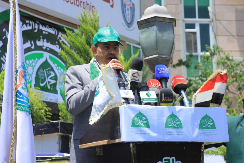 Man in a gray suit and green cap speaks into a cluster of microphones at an outdoor press conference, behind banners in Arabic and a lamppost.