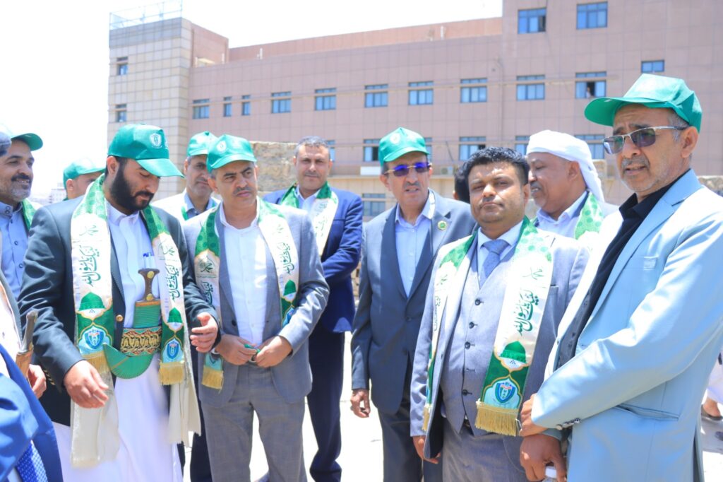Group of men in suits wearing green ceremonial stoles and hats, gathered outdoors for a formal event in front of a building.