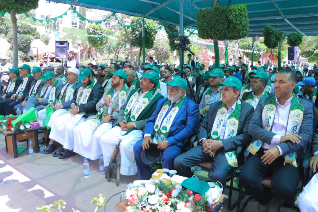 Group of men and women in green caps and sashes seated at an outdoor ceremony under a teal canopy, with plants and decorations in the background.