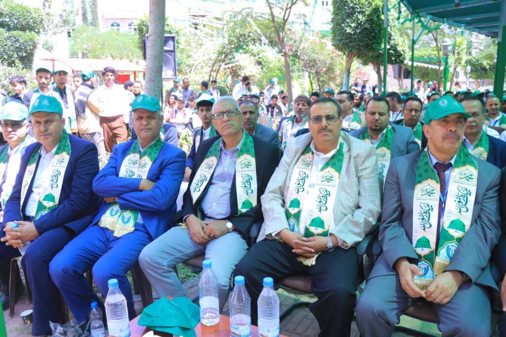 Group of men in suits seated outdoors at a ceremony, wearing green patterned sashes with logos and turquoise caps.