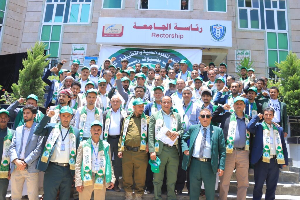Large group of men in green caps and scarves posing on steps in front of a university building with a Rectorship sign and a banner behind them, celebrating with raised fists.