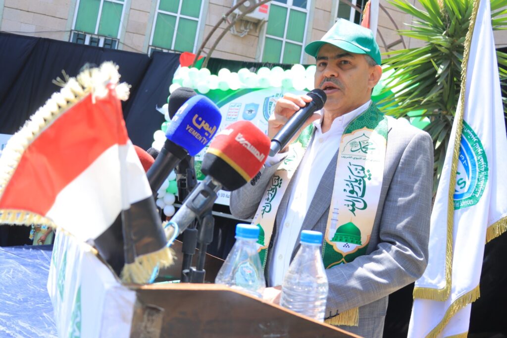 Man in a gray suit and green cap delivers a speech at an outdoor ceremony, wearing a decorative sash with green motifs.