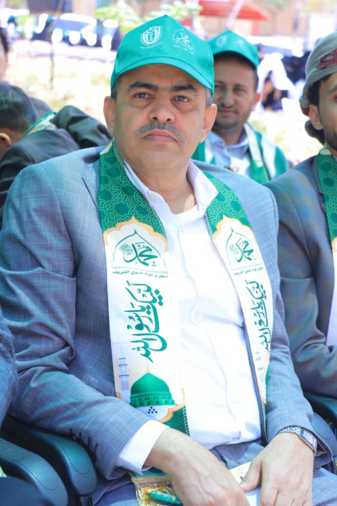 Man in a gray suit with a white shirt wears a teal cap and a green ceremonial scarf with Arabic text, seated at an outdoor event alongside others.